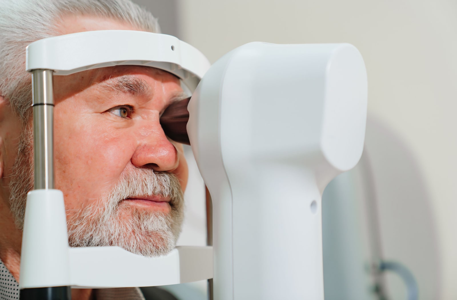A senior man is undergoing a corneal topography exam. His left eye is placed in a device while his chin is resting in a chin support from the machine.