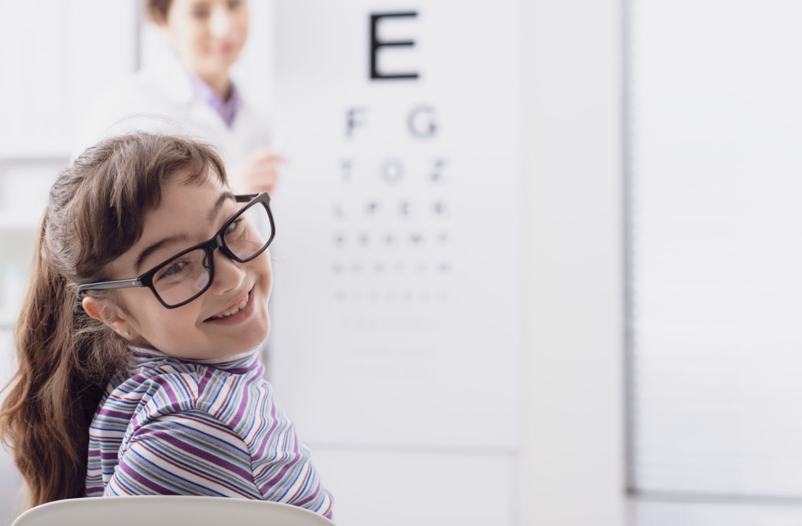 A young girl wearing glasses, leaning back and smiling towards the camera while she's in the optometrist's office