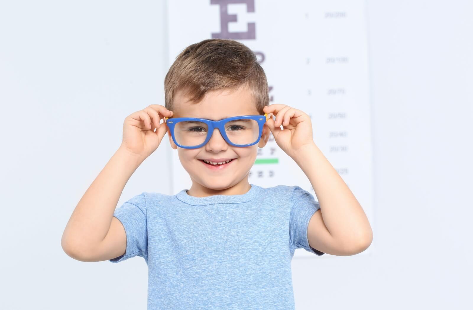 A little boy smiling in front of an eye chart while trying on myopia control eyeglass lenses.