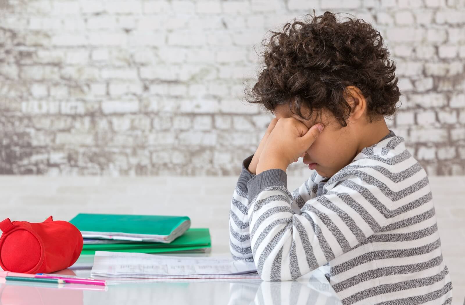 A child sitting with homework on a table rubbing their eyes with both hands.