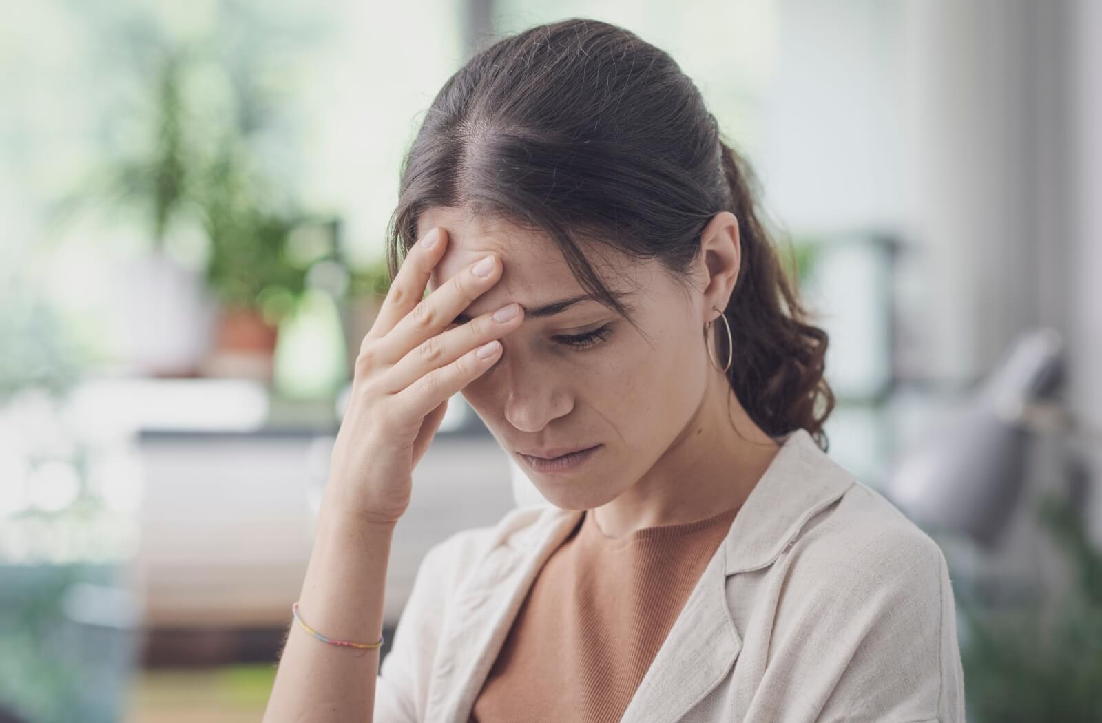 A person in a bright room grimaces in pain from a headache and rubs their face with their hands.