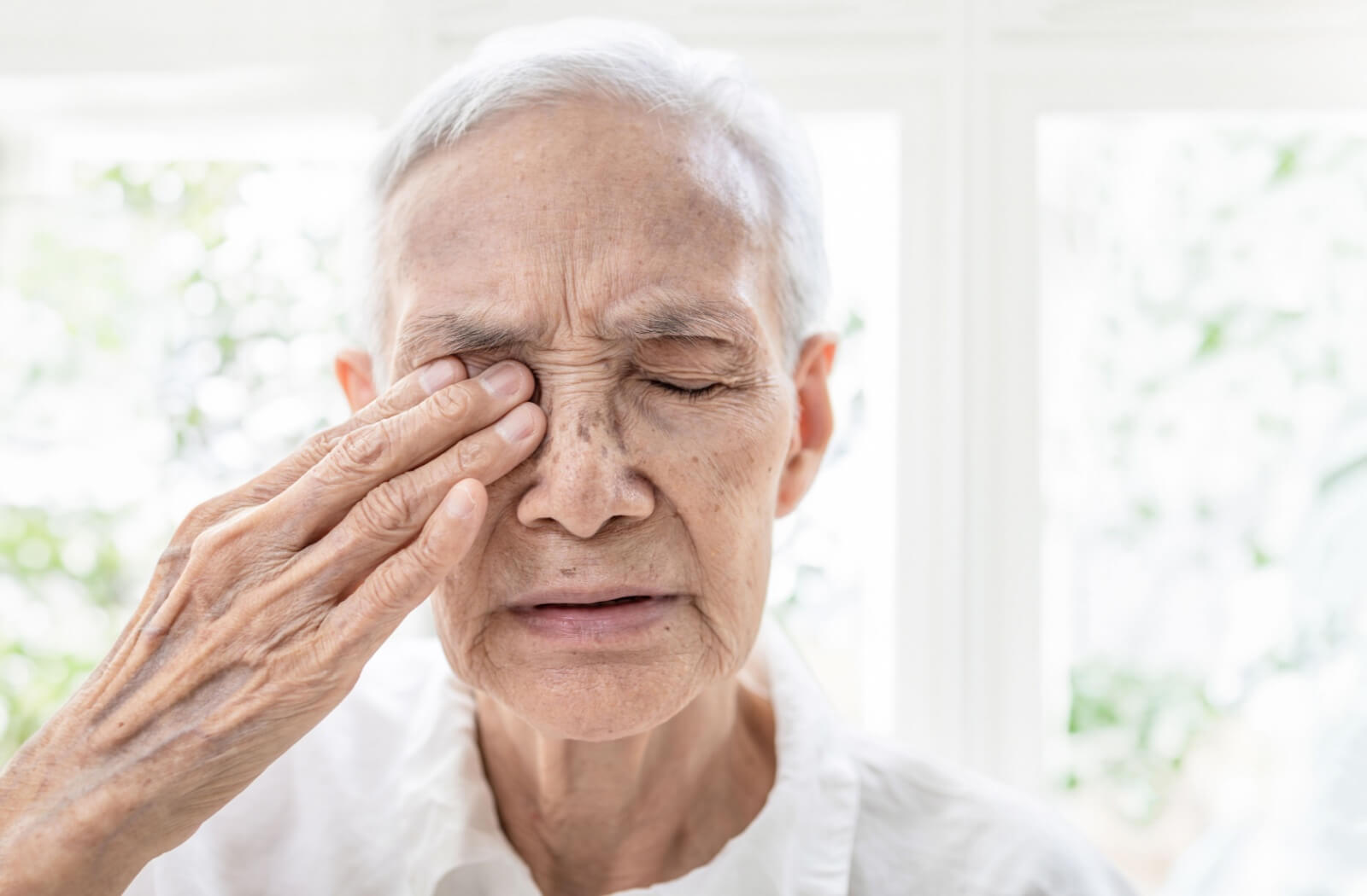 Older person with eyes closed, gently touching one eye, appearing to experience discomfort.