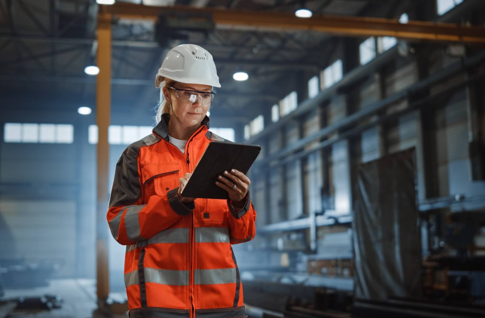 A person wearing a hard hat, prescription safety glasses, and a jacket uses a tablet while standing in a warehouse.