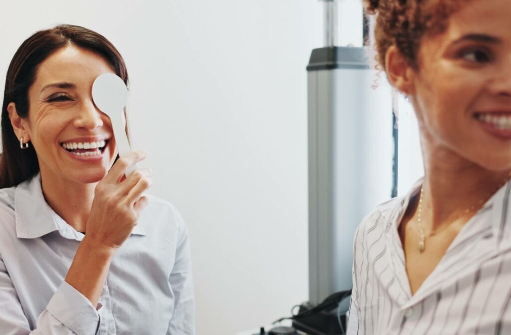 Smiling senior person holding an occluder over one eye during a vision test, while another person stands nearby, suggesting a friendly and relaxed eye exam environment.