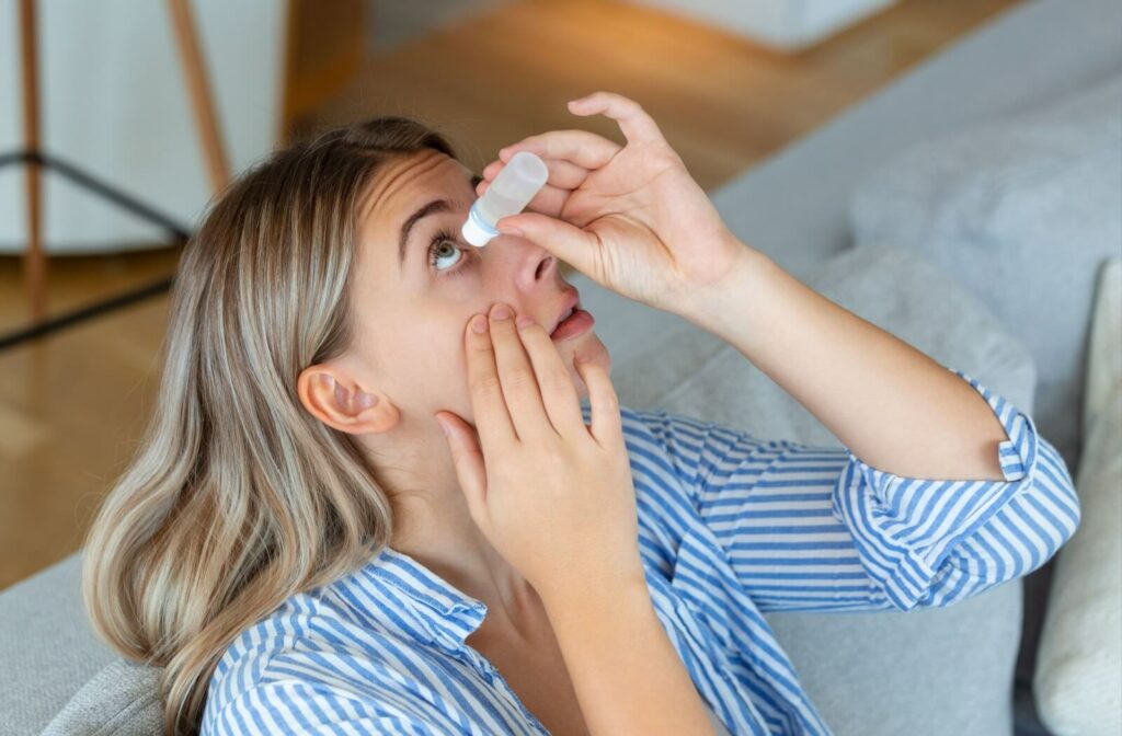 A person tilting her head back and applying eye drops while sitting on a couch, demonstrating proper use of lubricating or medicated drops for eye care.