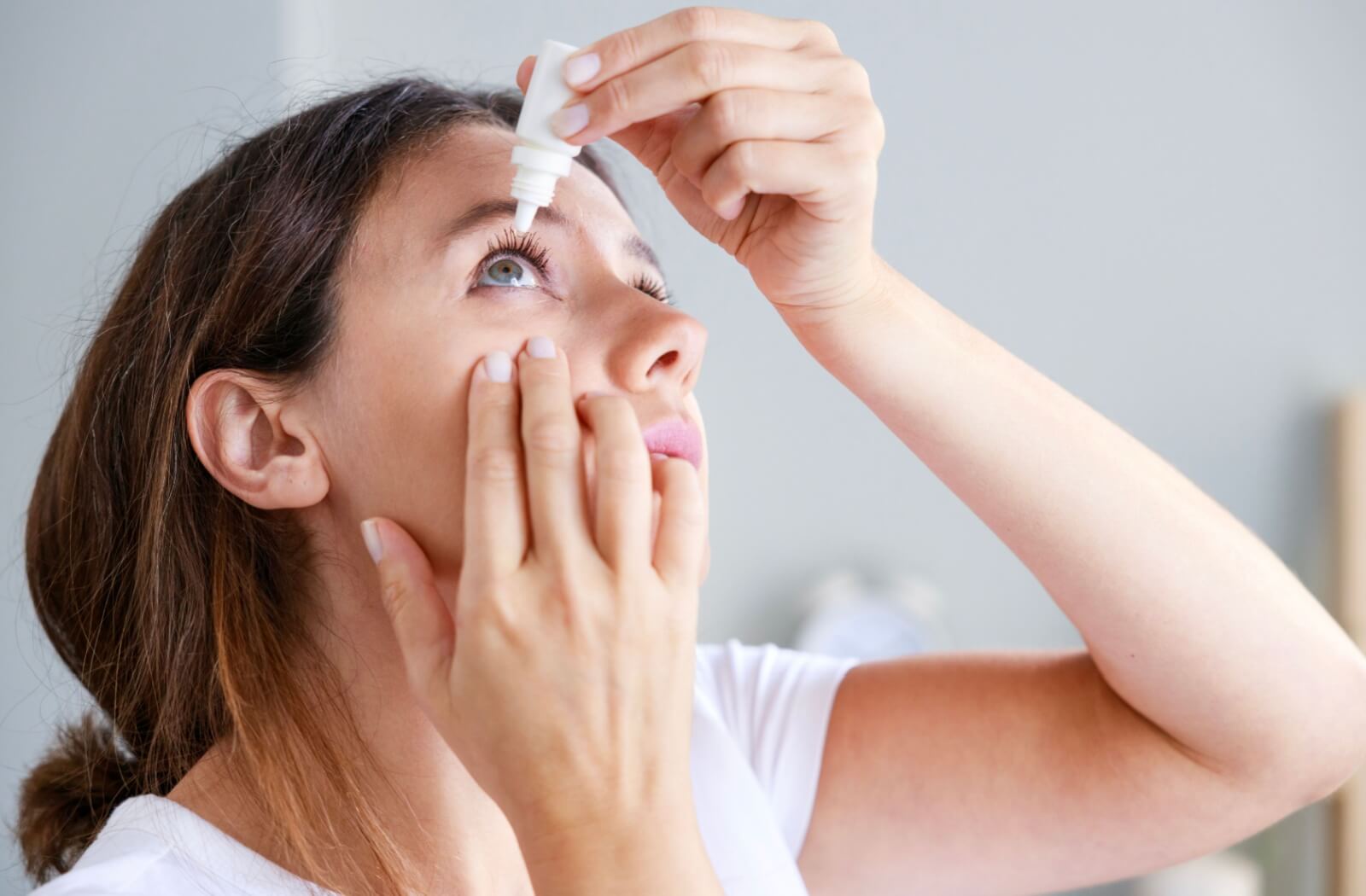 A woman applying eye drops to relieve her dry eyes, holding the bottle above her eye while gently pulling down her lower eyelid. The background is neutral, with natural lighting highlighting her actions.